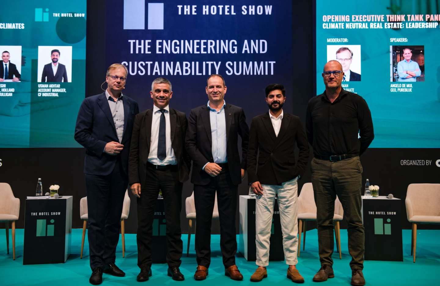 a picture of 5 men in suits standing at a conference together posing for a group picture