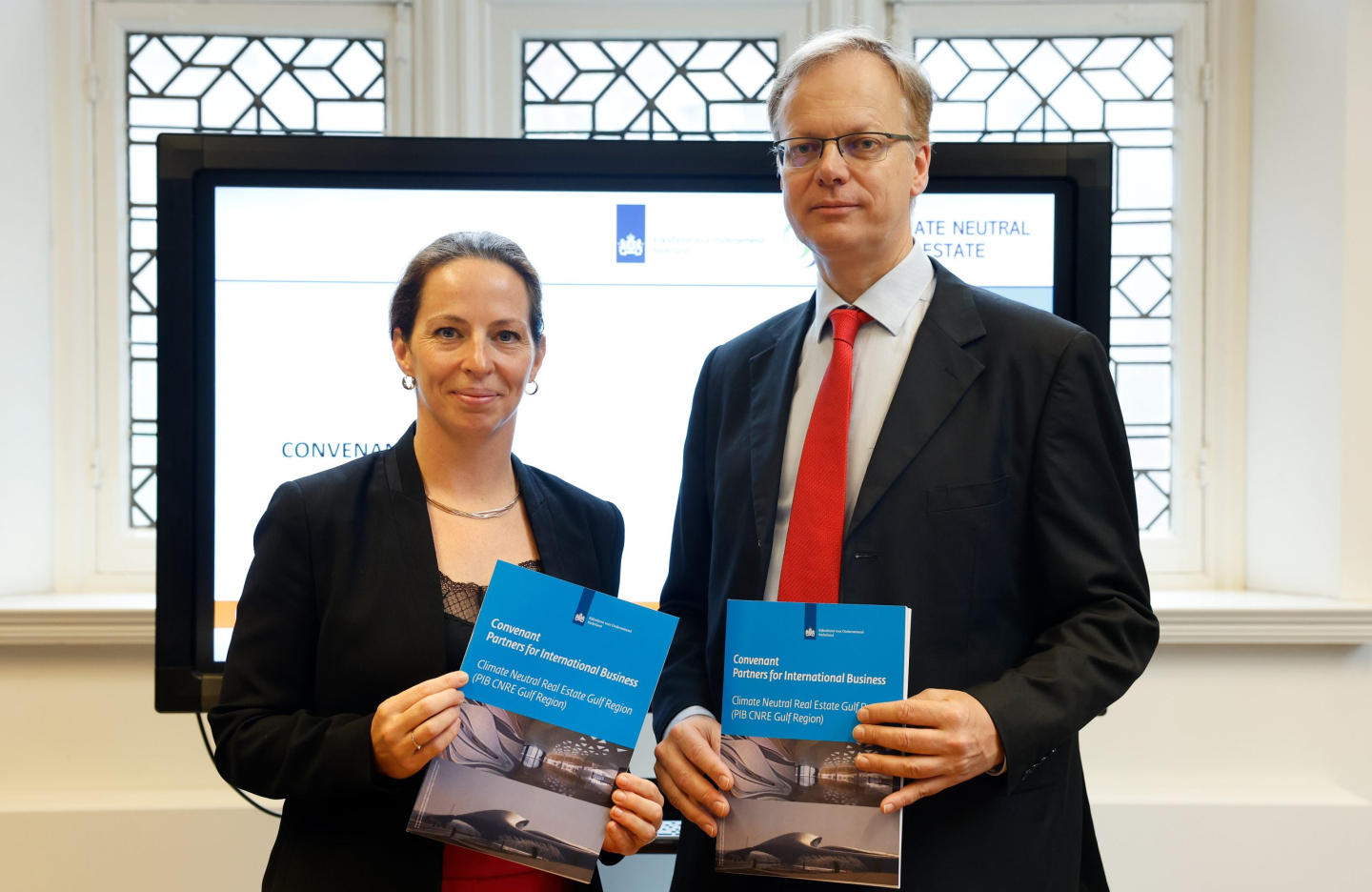 a man and a woman in suits pose with a folder featuring branding related to the Dutch Government