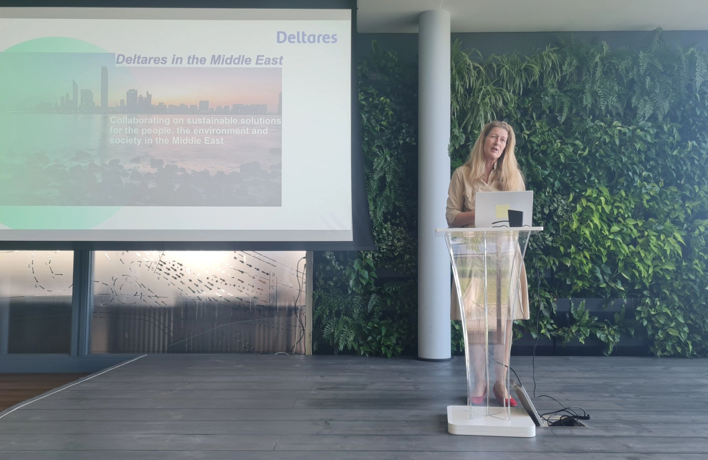 A lady stands at a conference easel on stage and presents with a powerpoint behind her on a screen