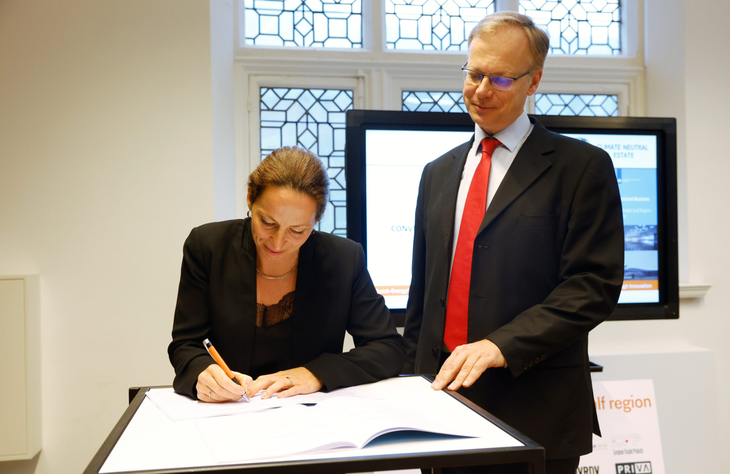 a woman in a blazer signs a document as a man in a suit and red tie stands next to her