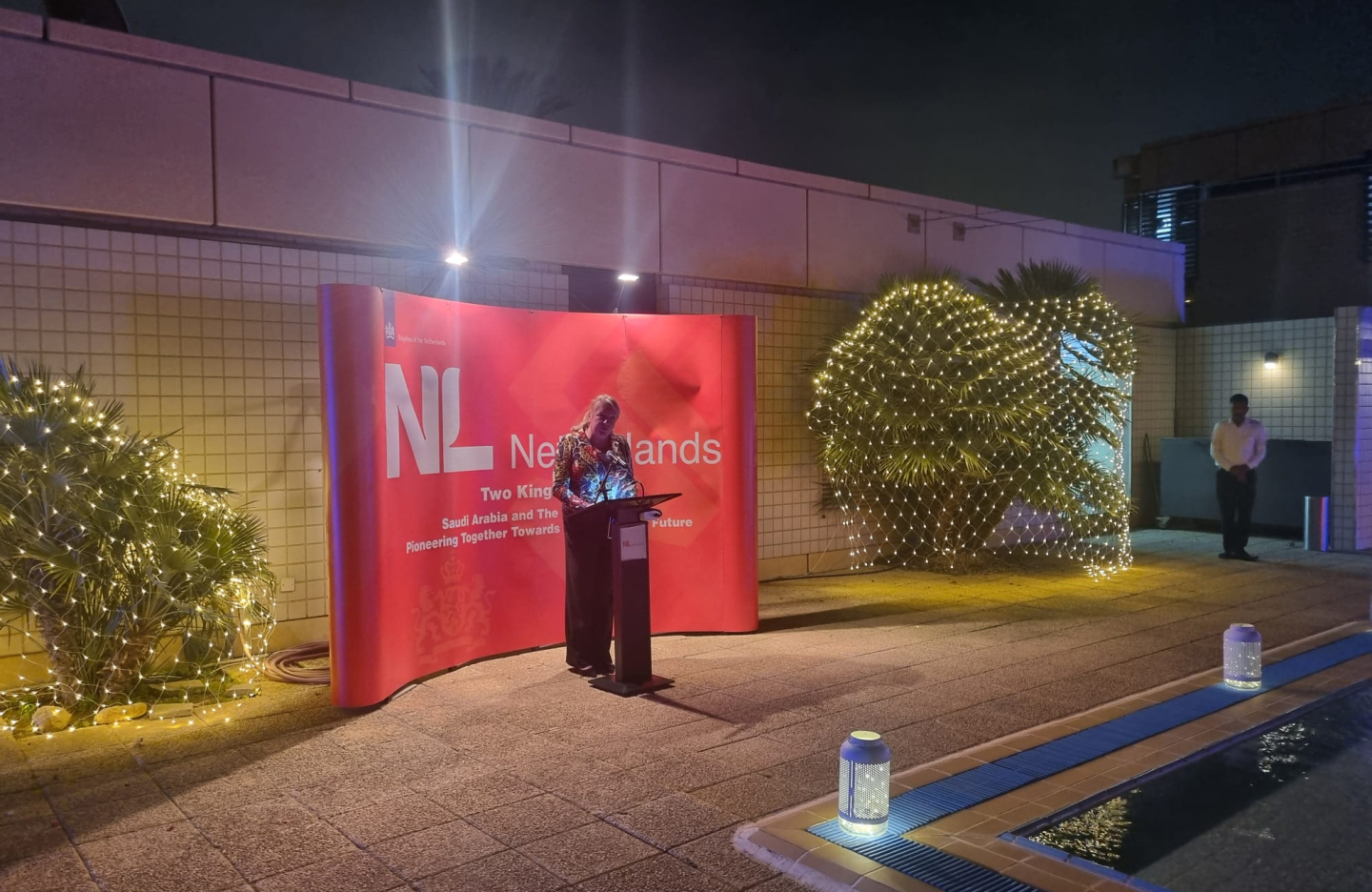 a woman presents on stage with a large orange backdrop and the NL Netherlands branding behind her