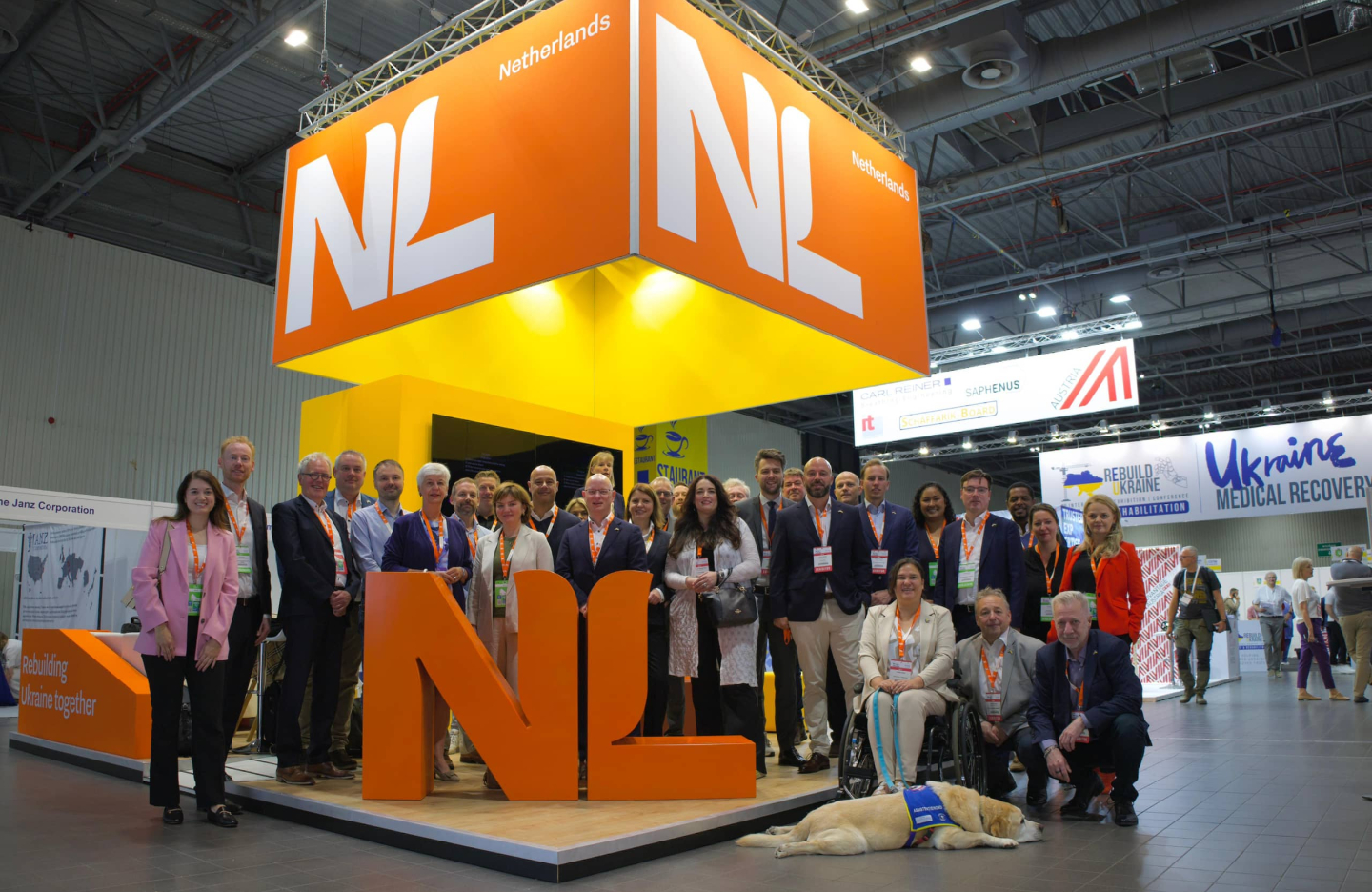 A group of participants in the ReBuild Ukraine 2025 mission take a group picture at the NL Pavilion. The large orange NL letters stand before them.