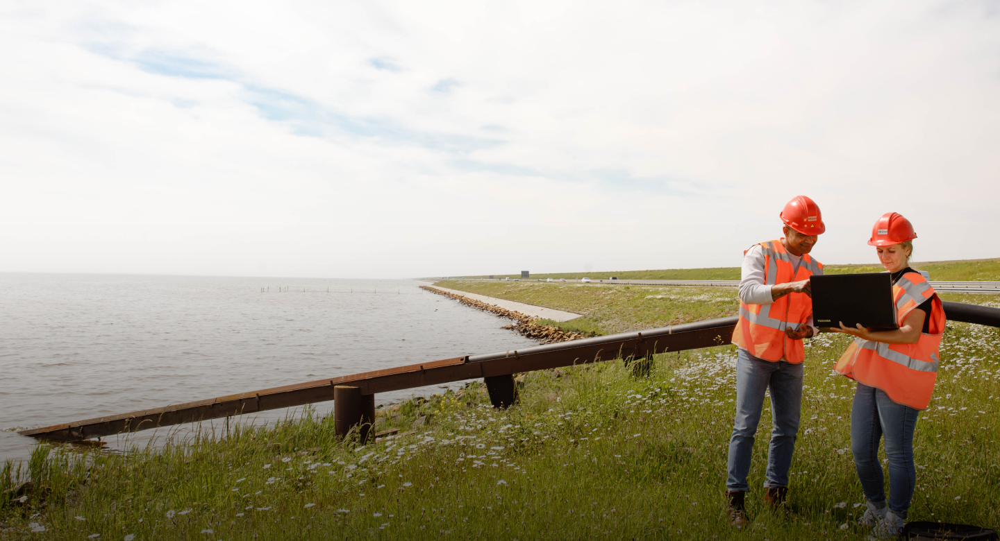 Two construction workers in high-vis vests and helmets stand next to a water body on green grass whilst looking at a clipboard 