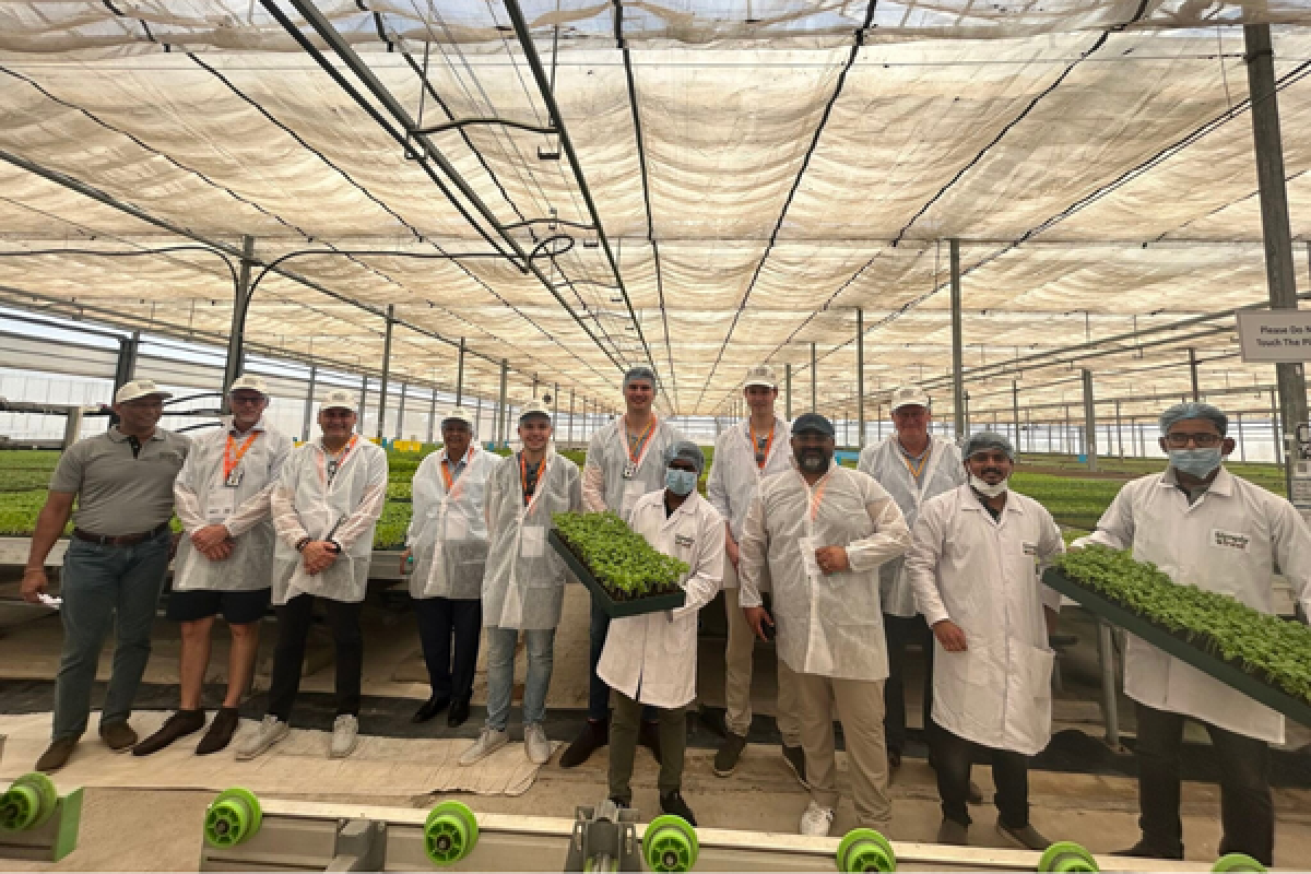 people in white lab coats proudly display crops by holding them up for the camera. Behind them is a large greenhouse.