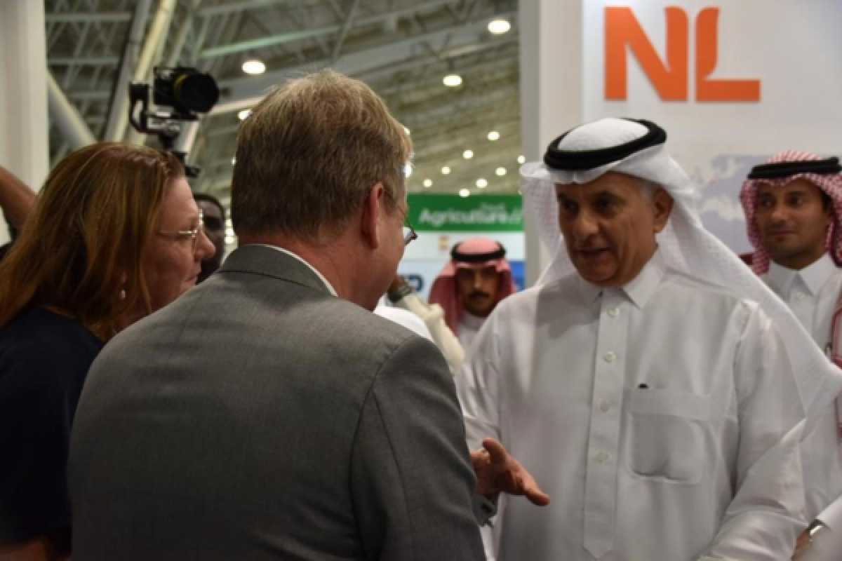 A man in traditional Emirati clothing shakes the hand of a Dutch man in a suit at a conference. In the background the orange NL letters are visible
